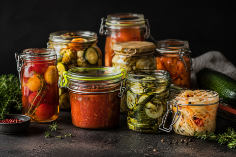 Plusieurs bocaux de conserves maison remplis de légumes et de sauces de saison sur un plan de travail sombre