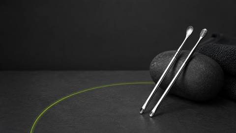 Un set de cure-oreilles oriculi en acier inoxydable poli argenté.posé sur une surface sombre texturée. Fond uni gris anthracite très foncé. Ambiance salle de bain minimaliste et premium, zéro déchet.
