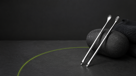 Un set de cure-oreilles oriculi en acier inoxydable poli argenté.posé sur une surface sombre texturée. Fond uni gris anthracite très foncé. Ambiance salle de bain minimaliste et premium, zéro déchet.