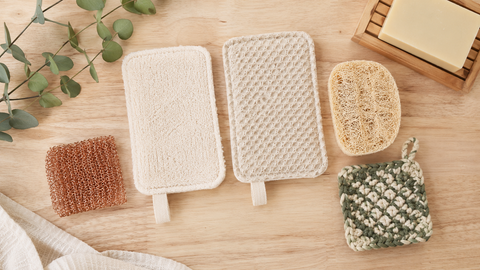 Un plan de travail en bois clair, Disposés harmonieusement : une éponge lavable en tiss, une tranche de loofah beige fibreux, un tampon récurant en fils de cuivre et un petit tawashi carré. 