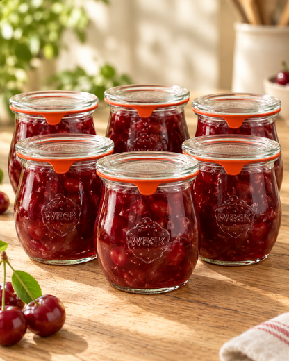 Lot de six bocaux Weck remplis de confiture de cerises maison, fermés avec leurs couvercles en verre et joints orange.